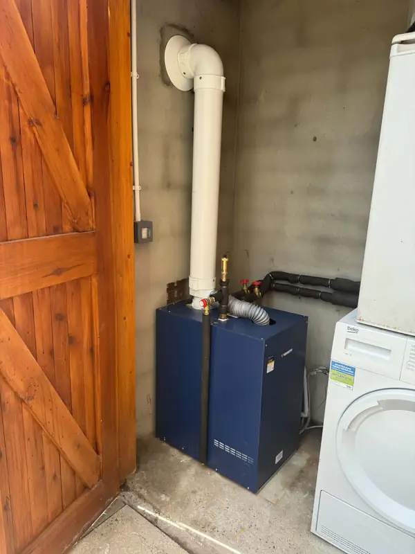 Blue Warmflow boiler fitted in an outhouse beside a washing machine, with a timber door