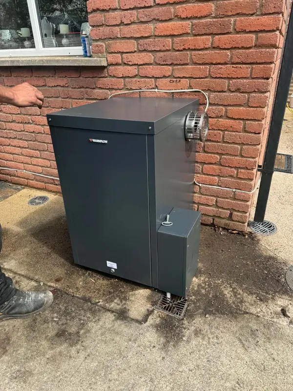 An SOS engineer's hand and boot beside an outdoor Warmflow oil boiler against a brick wall